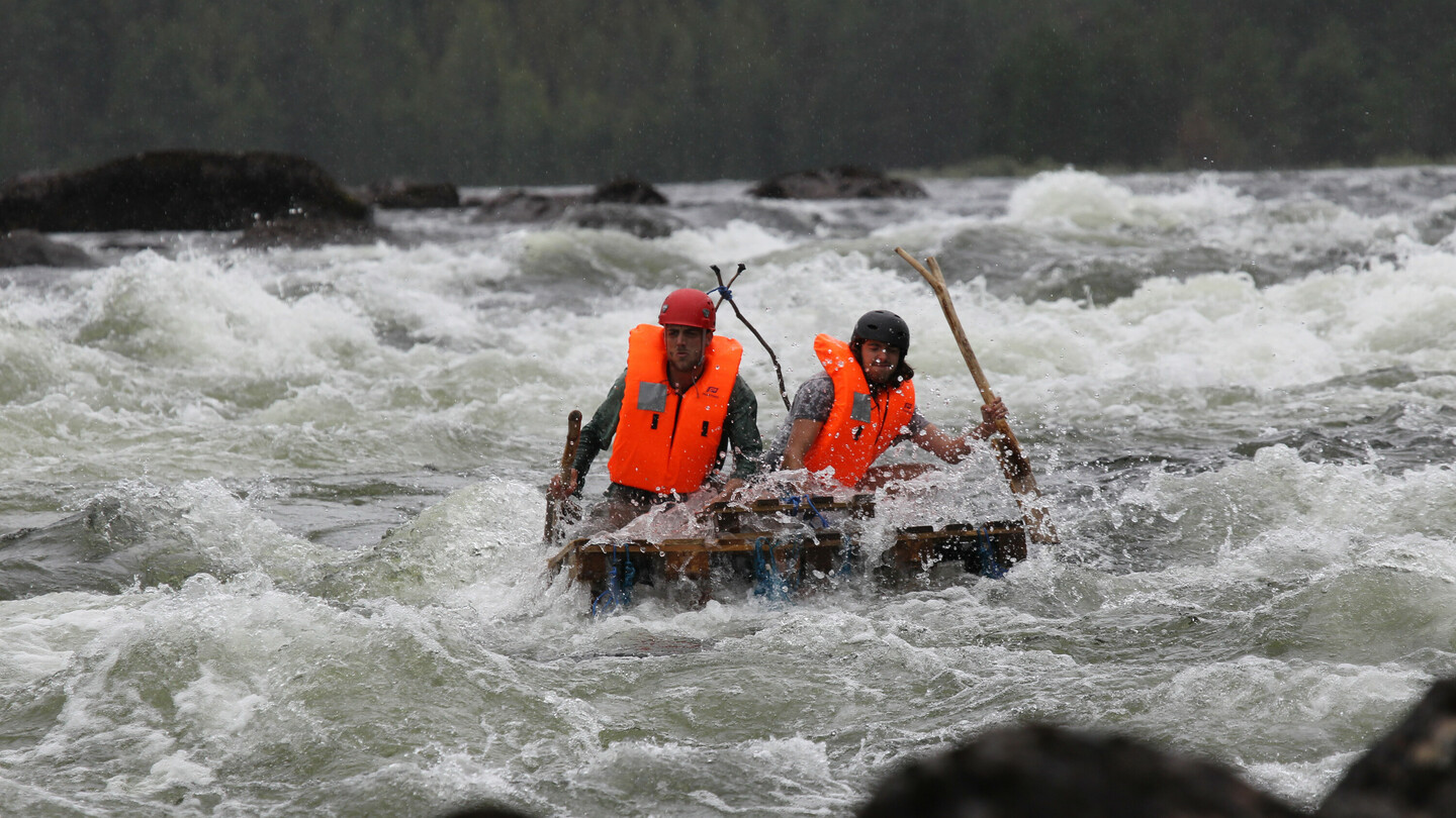 Two people wearing helmets and bright orange life vests navigate with paddles on a makeshift raft through turbulent, foaming rapids.